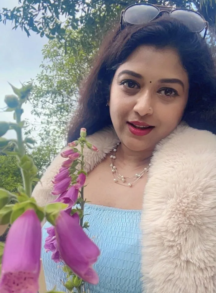 Shatabdi Mandal smiling in a garden, wearing a soft cream fur collar and pearls, with pink foxgloves in front of her